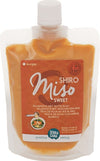 A stand-up pouch of TerraSana Shiro Miso Sweet paste with an orange matte label, white screw cap at the top, clear plastic packaging showing beige miso paste inside. The front label displays legible product name and ingredients in multiple languages, a simple illustrated bowl of soup, and block sans-serif typography. Product is photographed against a plain background with no people visible.