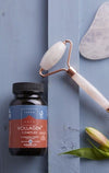 A jar labeled Vollagen Complex sits on a blue surface next to a jade roller and a small flower, highlighting a wellness or skincare routine. The jar is cylindrical with a white lid and matte light-colored body. Typography on the label is sans-serif. The jade roller has gold metal hardware and green stone ends. Small pale flower rests nearby. No people or hands are visible in this close-up product shot.