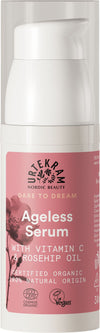 A bottle of Urtekram Ageless Serum with Vitamin C, Rosehip Oil, and vegan ingredients. The label highlights that it is certified organic and made from 100% natural origin. The packaging features a pink floral design. The bottle is cylindrical with a matte finish and pump dispenser. Label text uses clear sans-serif font in white and silver on a pink background. No people or hands are visible, plain backdrop, centered product shot.