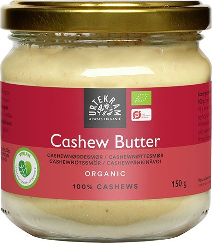 Urtekram Luomu Cashewpähkinävoi; lasipurkki Urtekramin luomu-cashewvoita varten punaisella paperietiketillä ja metallikannella; lieriömäinen purkki sisältää vaaleanbeigeä levitettä; etiketissä vegaani- ja luomusertifiointilogot, tuotenimi, teksti 100 % cashewpähkinöitä, paino 150 g ja monikielinen teksti; etiketin fontit groteskia normaalina, lihavoituna ja kursiivina; ei ihmisiä tai käsiä; neutraali tausta, keskitetty tuotekuva.