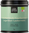 A black tin labeled Urtekram Always Organic contains whole bay leaves, featuring a green and beige design. The surface of the tin is matte. It is rectangular with rounded edges and a fitted lid. Sans-serif bold white text lists product details, including multiple languages and net weight of 5 grams, on the front label. The background is plain white with no people or hands visible in the shot.