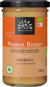 Urtekram Luomu Maapähkinävoi Crunchy; purkki Urtekramin luomu crunchy-maapähkinävoita oranssilla etiketillä, esillä ei lisättyä sokeria ja runsaasti proteiinia; vegaaniystävällinen levite 230 gramman purkissa mustalla kannella; purkki lieriömäinen, kirkasta lasia kiiltäväpinnalla; muovinen kansi mattamusta ja uurteinen; oranssissa paperietiketissä valkoista lihavoitua groteskitekstiä ja mustia yksityiskohtia; tuote yksin valkoisella taustalla studiokuvassa.