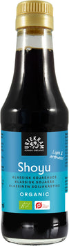 A bottle of Urtekram organic Shoyu soy sauce with a blue label, gold cap, and product information written in multiple languages. The bottle is dark-colored glass with a glossy finish and cylindrical shape. Label uses sans-serif font for brand name and product type. No people or hands present. Shot against a plain background with the entire bottle visible.