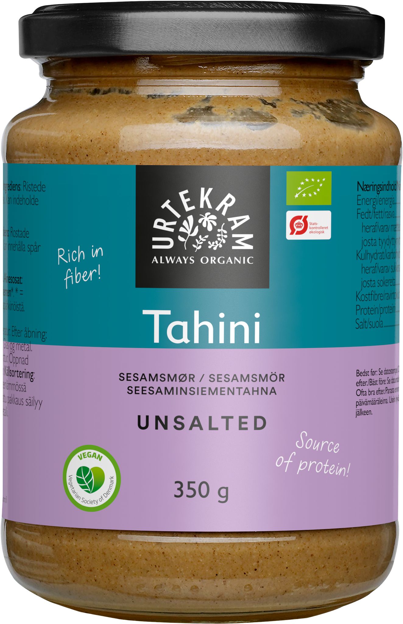 A cylindrical glass jar of Urtekram organic unsalted tahini with a purple matte label, black plastic screw cap, and clear body showing light beige contents. The label includes bold white and green text: “Rich in fiber!” and “Source of protein!”, along with product details and nutritional information in several languages. No people or actions visible; image is shot against a plain background. A jar of Urtekram organic unsalted tahini with a purple label, featuring Rich in fiber! and Source of protein! The