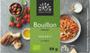 A package of Urtekram organic umami bouillon cubes is shown alongside bread, olive oil in a glass bottle, red cherry tomatoes, a small heap of salt, and a bowl of pasta on a smooth gray tabletop. The bouillon box is rectangular with matte finish and green color scheme; legible label reads “Urtekram” and describes the product. No people or hands are visible in this overhead shot.