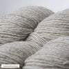 Close-up of light gray yarn skeins showing their soft, woolly texture. Material is loosely spun wool in a matte finish. Each skein is cylindrical with visible strands. The background is plain and light-colored. There are no people or hands visible. A small printed label with simple type in the corner reads “vaaleanharmaa.”.