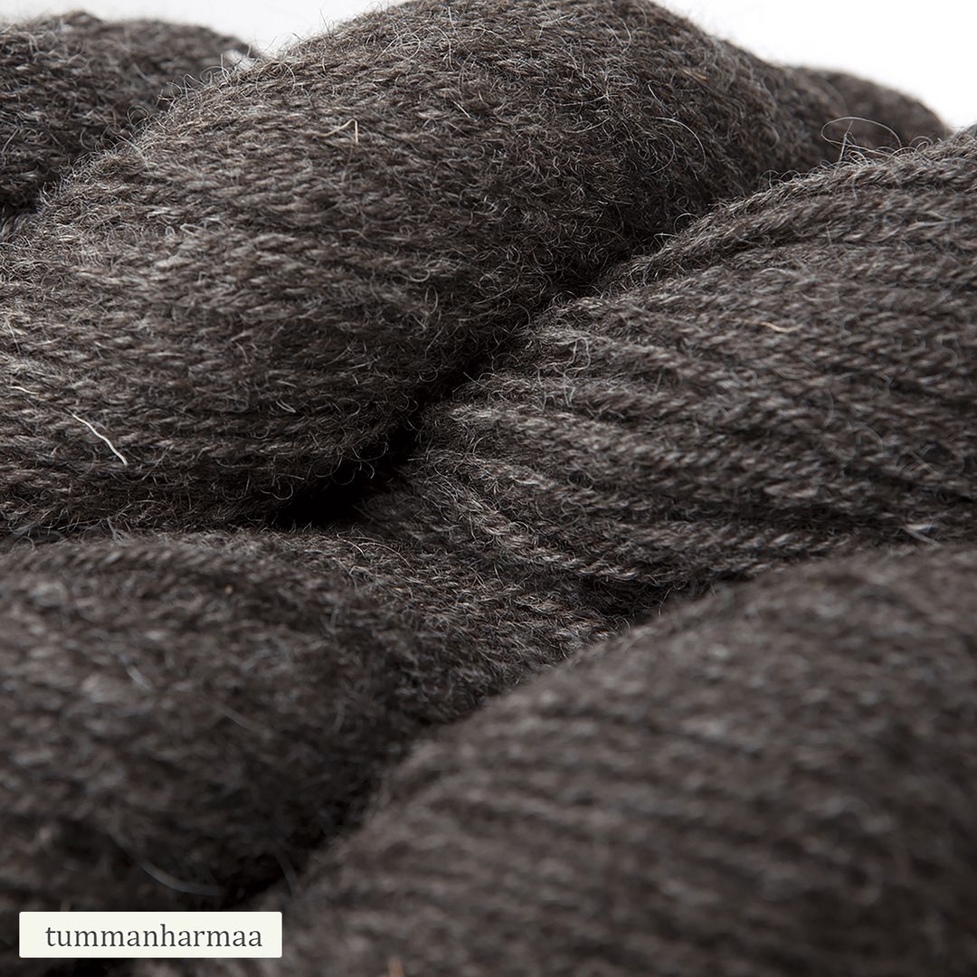 Close-up of thick, dark gray yarn skeins with a soft, slightly fuzzy texture. Multiple rounded skeins are tightly wound with visible fibers. Surface is matte. Yarn appears unwrapped and not in commercial packaging. A small, unobtrusive label in the corner has simple sans-serif text reading tummanharmaa. Plain background. No people or hands present.