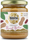 A jar of Biona Organic Peanut Butter, smooth and unsalted, 250g. Cylindrical glass jar with a wide gold metal lid and matte label. The label is dark with colored illustrations of peanuts and green leaves. Main text is bold, white sans-serif; smaller supporting text present. “Palm oil free” and “organic” are noted. Neutral plain background, no people or hands visible. Straight-on product shot.
