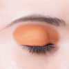 A close-up of a closed eye with shimmering orange eyeshadow applied to the eyelid. The finish of the eyeshadow is metallic and reflective. The eyebrow above is well-groomed with defined edges, and individual dark eyelashes are visible along the eyelid margin. No product packaging, label text, or additional items are present in the image.