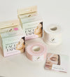 Two rolls of FACEIMAGE kinesiology face tape, one pink and one white, are displayed with their boxes and an instruction booklet featuring a woman’s face with white kinesiology tape on her cheeks and forehead. Rolls have visible perforation lines; tape has a matte finish. Packaging is rectangular with product name in bold sans-serif font. Booklet cover is mostly white; photo background is plain and light-colored.