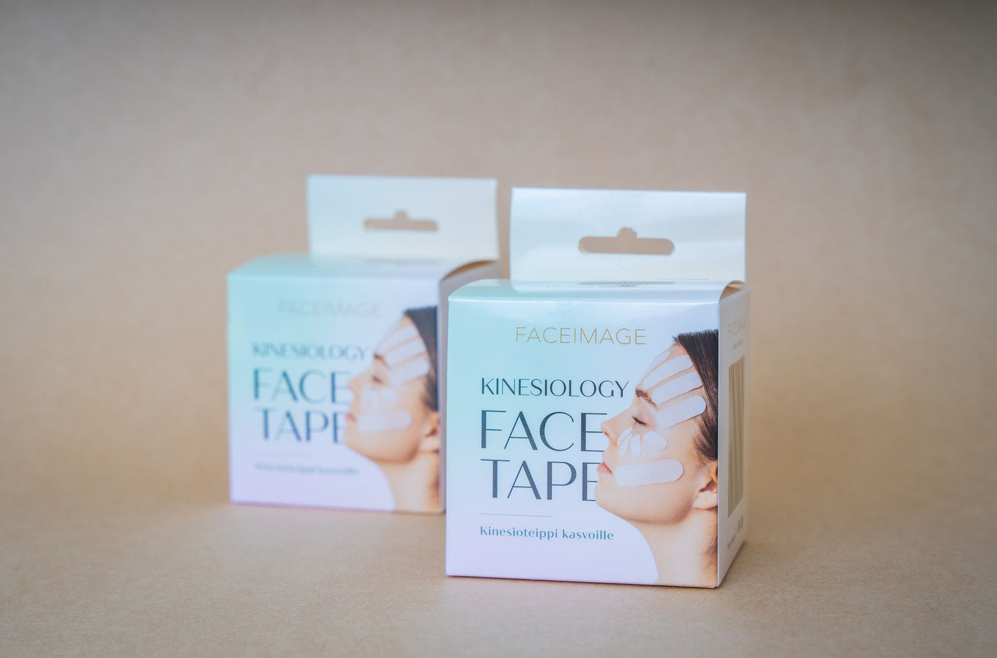 Two boxes of Faceimage Kinesiology Face Tape are shown against a beige background. Boxes are rectangular with a matte finish, primarily white and beige in color. Each features a close-up portrait of a woman with pink tape applied to her cheeks, forehead, and chin. The product name on the box is in bold sans-serif font at the top. No hands or additional objects visible.