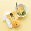 A bowl of pasta with greens and grated nutritional yeast sits on a yellow surface beside a white napkin and a yellow bag labeled Ravintohiiva Hiutale. A spoon is in the bowl. The yellow bag is matte, rectangular, and displays bold, black sans-serif text on the front. The background is evenly lit, overhead view; no people or hands are present.