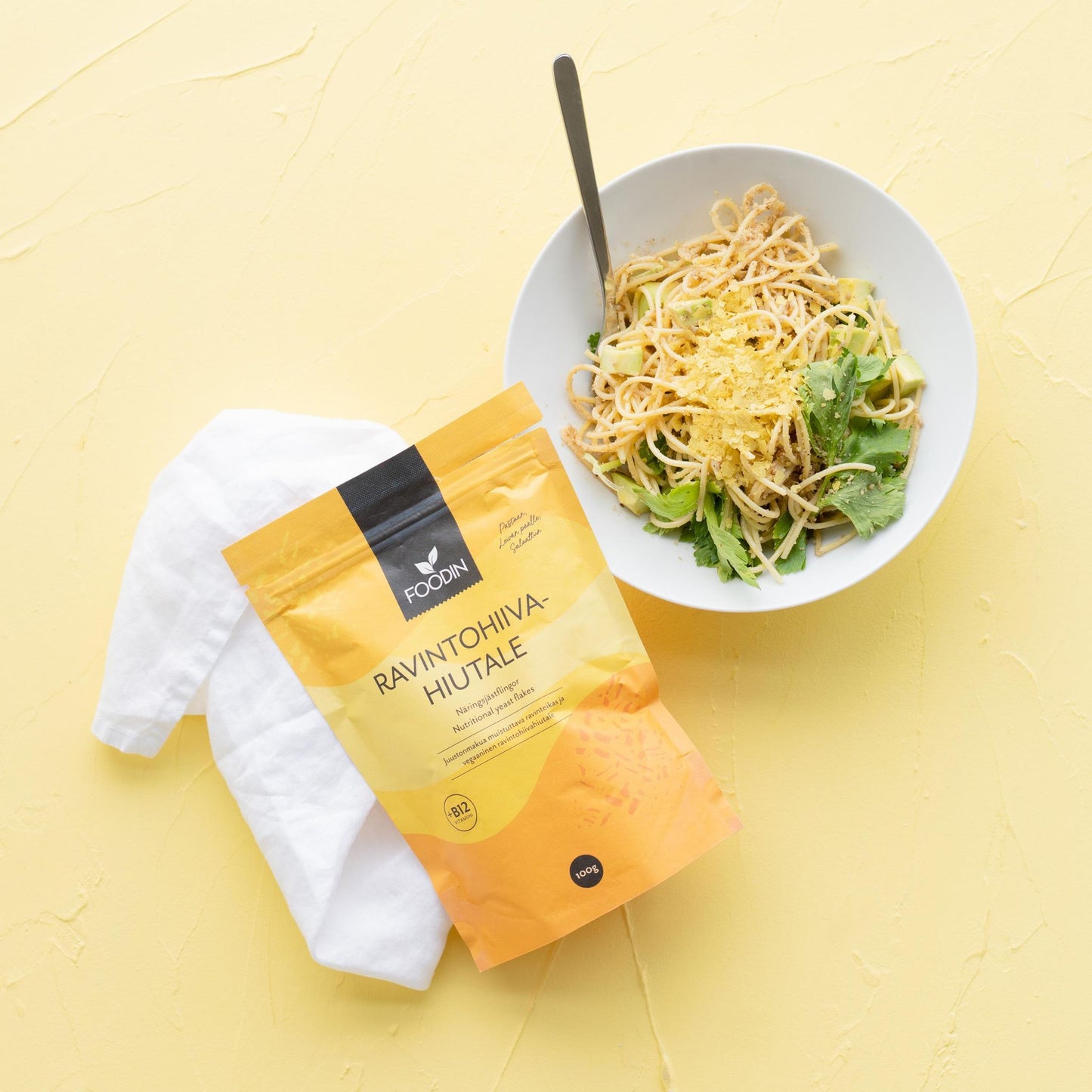 A bowl of pasta with greens and grated nutritional yeast sits on a yellow surface beside a white napkin and a yellow bag labeled Ravintohiiva Hiutale. A spoon is in the bowl. The yellow bag is matte, rectangular, and displays bold, black sans-serif text on the front. The background is evenly lit, overhead view; no people or hands are present.