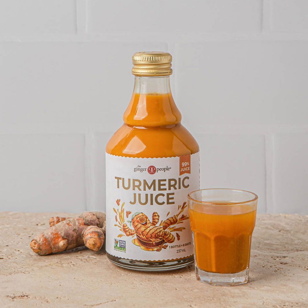The Ginger People kurkumamehu; pullo tekstillä "Turmeric Juice" pystyssä samaa kirkkaan keltaisenoranssia nestettä sisältävän lasin vieressä; useita kokonaisia tuoreita kurkumajuuria vierellä; sileä, vaalea kivipinta.