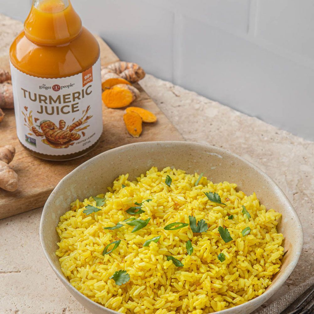 The Ginger People kurkumamehu; kulhollinen eloisan keltaista kurkumariisiä vihreällä yrttikoristelulla kirkkaan, kullankeltaista nestettä sisältävän kurkumamehupullon vieressä puulaudalla, jolla myös viipaloitua raakaa oranssia kurkumajuurta; pullon etiketti yksinkertaisella groteskilla.