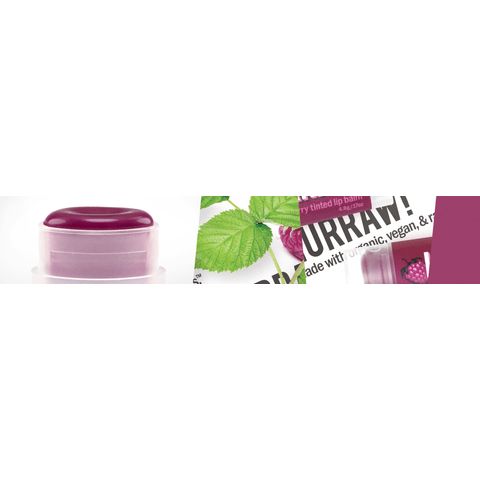 Hurraw! Sävyttävä Huulivoide Vadelma; pieni violetti huulivoidepurkki vastaavalla violetilla kannella, molemmat kiiltäväpintaisia, aseteltuna vihreiden lehtien ja violettien marjaryppäiden viereen; purkki pyöreä ja muovia; osittain näkyvissä painettua tekstiä groteskifontilla valkoisella taustalla; ei ihmisiä tai käsiä kuvassa.
