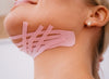 Woman with blonde hair and gold earring has pink kinesiology tape applied in strips under her chin and along her jawline, enhancing facial contouring. Tape is matte-finish, solid pink, cut into uniform narrow strips. No visible packaging or typography. Neutral background. Cropped close-up shot focused on lower face and neck; woman’s hands are not visible.