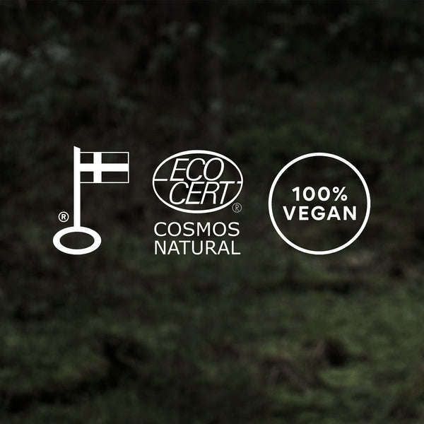 Taika Kosteuttava Käsivoide; kolme logoa tummalla taustalla: Pohjoismainen ympäristömerkki Joutsen, ECOCERT Cosmos Natural -sertifiointi ja "100% Vegan" -merkintä korostavat ekologisia ja luonnollisia tuotestandardeja; kaikki logot valkoisia selkein muodoin ja symbolein; tausta yhtenäisen musta, mattapinta; ei tuotteita, pakkauksia, ihmisiä tai esineitä kuvassa; lähikuva pelkistä logoista.