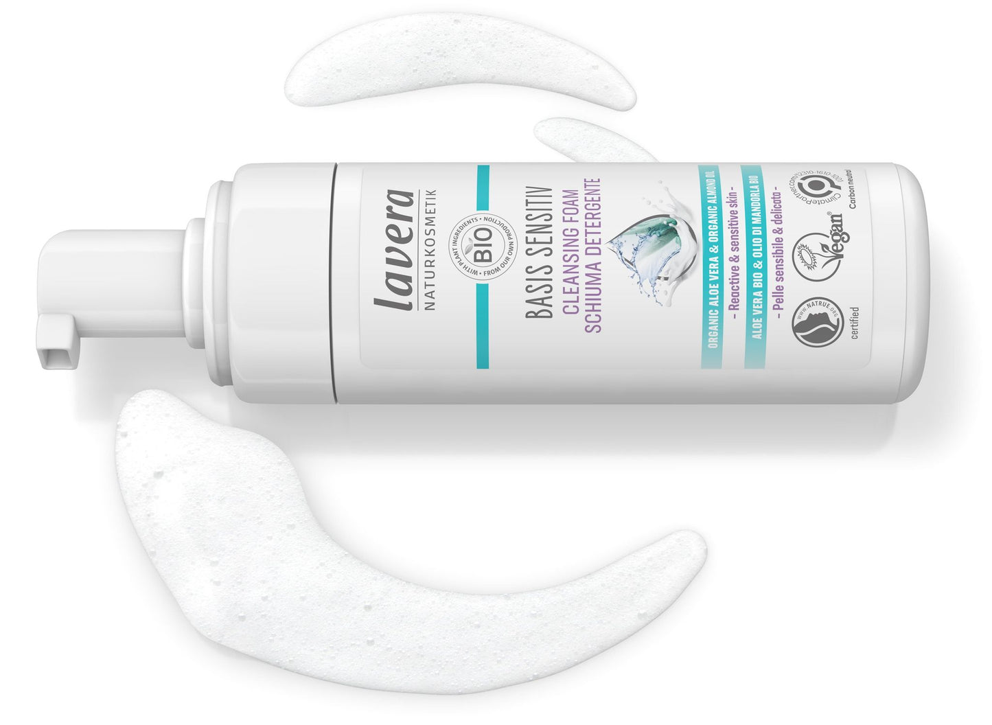 A plastic bottle of Lavera Basis Sensitiv Cleansing Foam with a white pump dispenser is positioned horizontally on a smooth, solid white surface. White foam is dispensed in curved formations near the bottle. The packaging is mainly white and blue with black sans-serif text and icons indicating organic, vegan certifications. No people or hands are visible in the image.