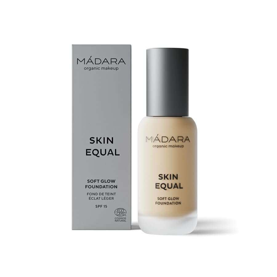 A clear cylindrical bottle with a pump dispenser and black cap containing liquid foundation stands upright beside a rectangular gray cardboard box. Both items display “MÁDARA Skin Equal Soft Glow Foundation SPF 15” in sans-serif lettering on the front. The scene has a plain, light-toned background with no people or hands visible.