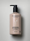 A beige bottle of Mádara Bitter Honey Moisture Wash with a black plastic pump is displayed on a smooth, light gray surface. The cylindrical bottle has a matte finish and features minimalistic white floral graphics. The label uses clean sans-serif typography, indicating it is for hands and body. No people or other objects are present in the shot.