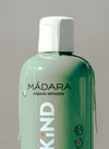 Madara KIND Mild Shampoo; lähikuva vihreästä, lieriön muotoisesta ja mattapintaisesta pullosta valkoisella muovikorkilla, etiketissä lihavoitu versaaliteksti "KIND" ja ekologisia kuvakkeita; hoitavaa nestettä valumassa pullon kylkiä pitkin; neutraali tausta.