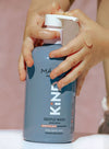 Child’s soapy hands pressing the pump of a matte blue liquid soap bottle with a white pump dispenser, labeled “KIND Gentle Wash” in bold white sans-serif lettering, on a solid soft pink background. Text on bottle notes suitability for sensitive skin and both kids and adults. Cropped close-up shot focuses on hands and bottle.
