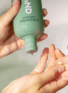 A person pours shampoo from a green MADARA bottle onto their hand, with a soft-focus background enhancing the soothing scene. The bottle is cylindrical with a matte green finish and features white uppercase sans-serif typography for the brand name “MADARA.” Only hands are visible, positioned in the foreground; no additional objects or label text are legible. The background is light-toned and out of focus.