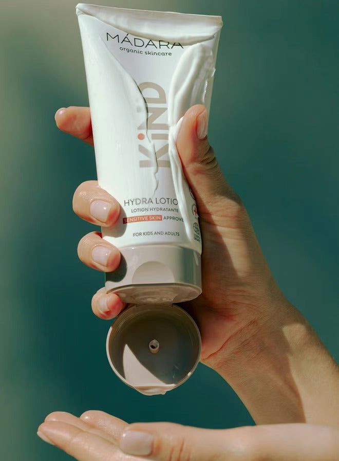 A hand squeezes Mádara organic skincare KIND hydra lotion onto another hand, with the cap open and a softly blurred background. The product is a cylindrical tube, pale peach-pink in color, with a matte finish and white sans-serif text labeling it as “KIND hydra lotion.” Tube cap is white plastic and hinged. Only hands are visible in the image.