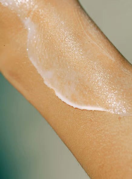 Madara KIND Hydra Lotion vartaloemulsio; lähikuva henkilön käsivarresta, jolle on levitetty leveä juova kosteuttavaa, puolikiiltävää valkoista voidetta antaen iholle kimaltavan ja kosteutetun ilmeen; ei näkyvää pakkausta tai tekstiä; tausta neutraali ja epätarkka.
