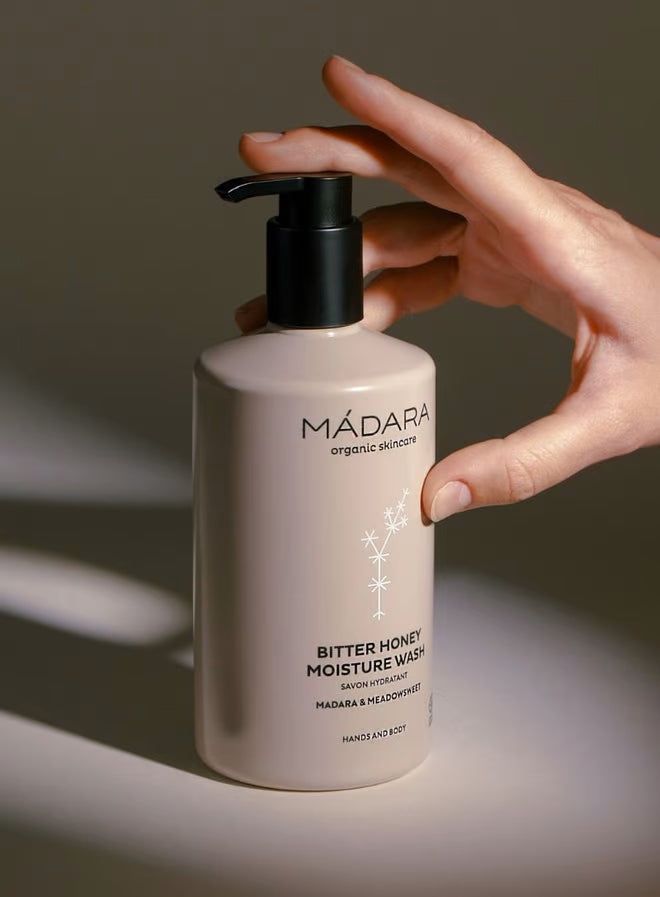 Madara BITTER HONEY kosteuttava saippua; käsi tarttuu beigeen, mattapintaiseen ja pyöristetyin reunoin varustettuun pumppupulloon, jonka etusivulla musta groteskiteksti "MÁDARA organic skincare, Bitter Honey Moisture Wash"; neutraali tausta, näkyvissä vain käsi ja kyynärvarsi.