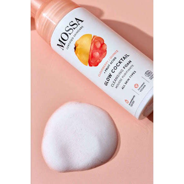Mossa Glow Cocktail Kirkastava puhdistusvaahto; sylinterinmuotoinen pullo Mossa Glow Cocktail Cleansing Foam -tuotetta valkoisella pumpulla, tasaisella persikanvärisellä pinnalla pienen valkoisen vaahtokasan vieressä; pakkaus kiiltäväpintainen, pääosin valkoinen värikkäin hedelmäkuvituksin ja pienin ihonhoitoaiheisin kuvakkein; teksti "Mossa Glow Cocktail Cleansing Foam" selvästi näkyvissä etiketissä; ei ihmisiä kuvassa.