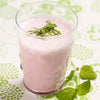 A glass of pink smoothie topped with shredded green herbs, placed on a patterned white and green surface with fresh mint leaves and vibrant herbs beside the glass. The smoothie is in a clear cylindrical glass without visible branding or labels. The surface has a repeating geometric pattern, combining white and green colors. No people or hands are present in the image.
