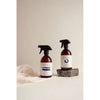 Two amber spray bottles labeled SEES stand on a light surface; one is placed on a small stone, while the other rests next to a white cloth. The bottles are cylindrical and have black spray tops with a matte finish. Labels display all-caps sans serif text. No people present. The background is plain off-white and uncluttered, with soft diffuse lighting.