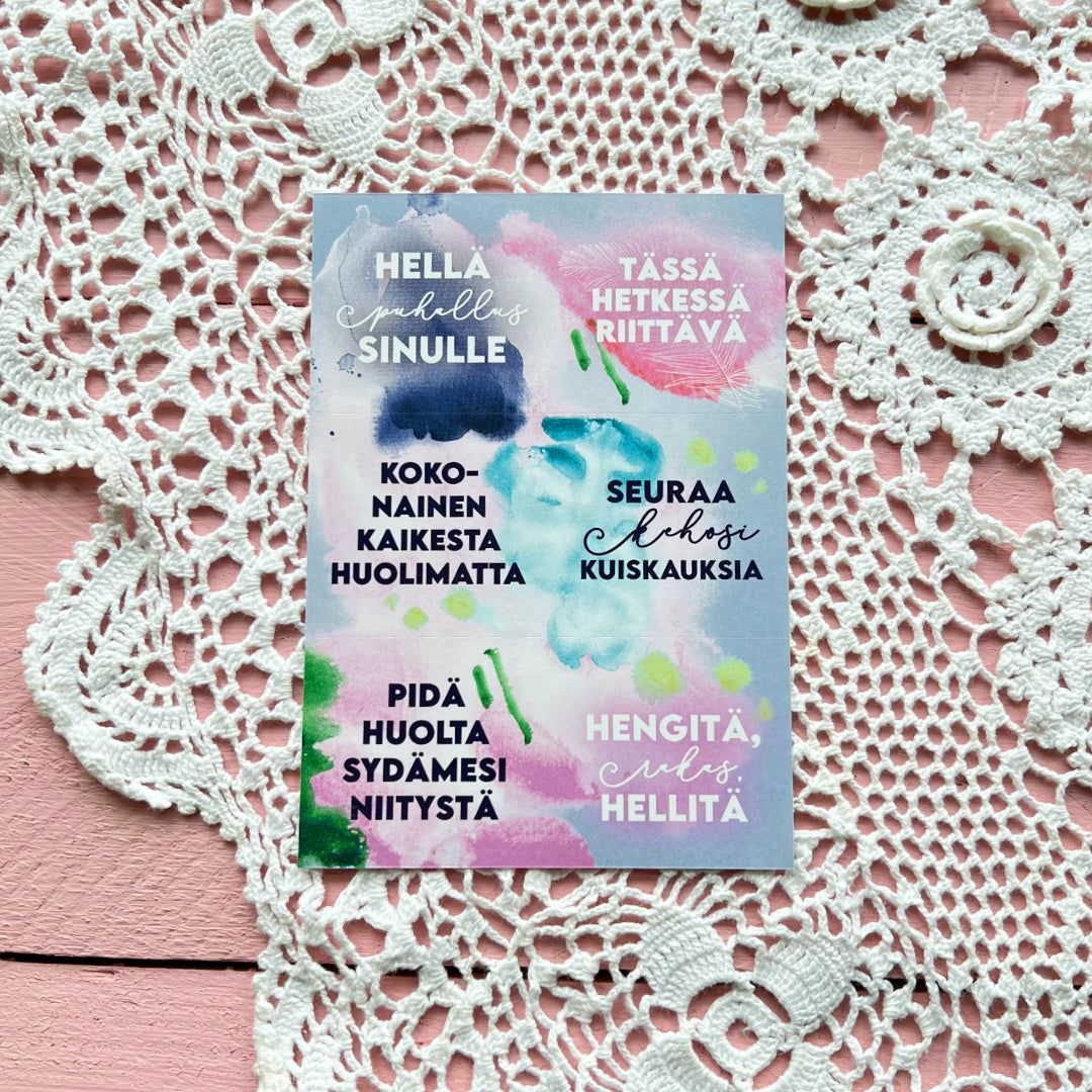 A postcard featuring Finnish motivational phrases on pastel watercolor splotches, displayed atop a white crochet doily and a pink wooden surface for inspiration. The postcard is rectangular and uncoated matte. Watercolor splotches include blue, green, yellow, and pink tones. Black sans-serif text in various weights is arranged diagonally and vertically within the colored splotches. No packaging or people are visible. Photographed from above in flat lay style.