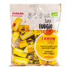 A clear bag of Super Fudgio banana fudges with yellow wrappers. Bag is plastic with matte finish, rectangular shape, and heat-sealed top. Front label uses bold sans-serif typography for the brand name with product details in smaller print. Vegan, gluten-free, dairy-free claims and coconut sugar/powder ingredient are stated. Nutritional info appears on one side of the pack. Background is plain white; no people or hands visible.