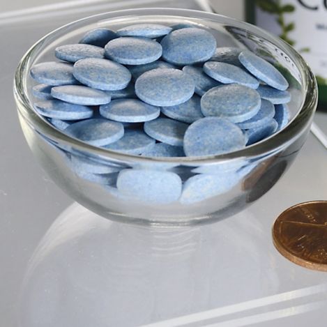 Swanson Copper 2 mg; kirkas lasikulho pyöreitä, litteitä sinisiä tabletteja kiiltävällä lasialustalla amerikkalaisen pennin vieressä kokovertailua varten; kuvattu ylhäältä; ei näkyvää pakkausta, etikettejä tai tekstiä; ei ihmisiä tai käsiä, sekava tausta.