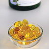 Swanson Vitamin E Mixed Tocopherols 400IU; pieni lasikulho kullankeltaisia vitamiinipehmeäkapseleita vaalealla alustalla, taustalla sumennettu pullo; soikeat, läpikuultavat ja kiiltäväpintaiset kapselit; sumennuksen vuoksi ei luettavaa etikettitekstiä; ei ihmisiä tai käsiä; kuvattu yläviistosta.