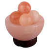 A pink Himalayan salt bowl holds four round massage stones in various light pink shades, resting on a dark round base. All items have a matte finish. No visible labels or packaging. Background is plain and neutral, and no people or hands are present. This is a close-up product shot focused directly above the items.
